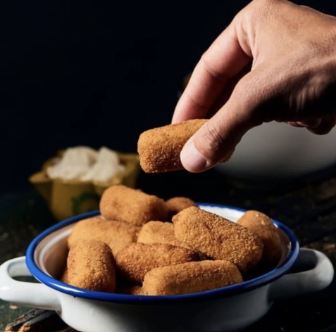 Main qui attrape une croqueta de jambon ibérique dorée dans un bol en céramique bleu et blanc