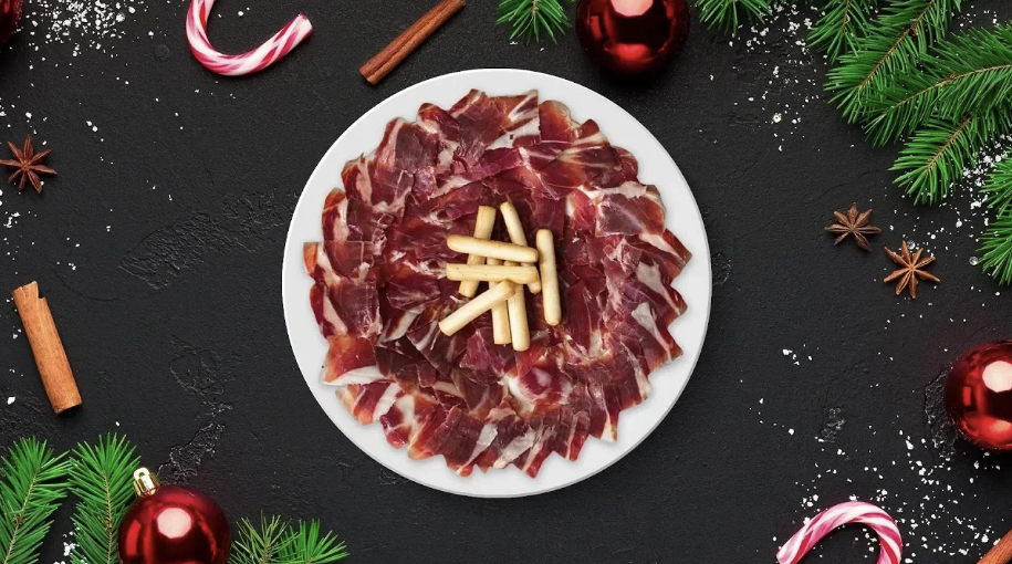 Assiette de jambon ibérique tranché avec gressins sur fond noir décoré de branches de sapin et boules de Noël